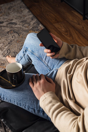 Attractive happy smart young man sitting on a floor in the living room, using phone, drinking coffeeの写真素材
