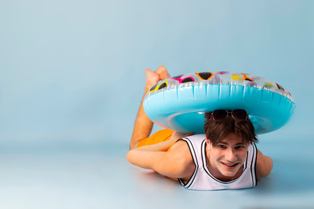 Funny man with blue inflatable swimming circle flaunts his strength on light blue background. Full length of playful strong male swimmer. Summer vacation concept.の写真素材