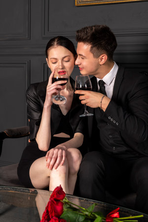 A young couple elegantly and stylishly dressed spend a romantic time with glasses of wine in hand while sitting on a vintage couch in a beautiful dark room in Gothic style. A romantic date.の写真素材