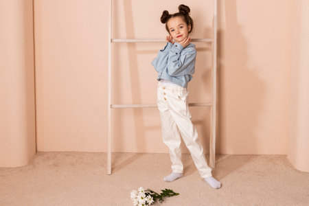 Portrait of a charming little girl, well dressed in a blue shirt and white pants or trousers and with a slight makeup and double bun hair. Isolated on pastel beige background.の写真素材