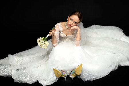 A beautiful charming girl, the bride in a luxurious and elegant white wedding dress, sadly and upset sitting on a floor with a graceful bouquet of white roses. Isolated on a black background.の写真素材