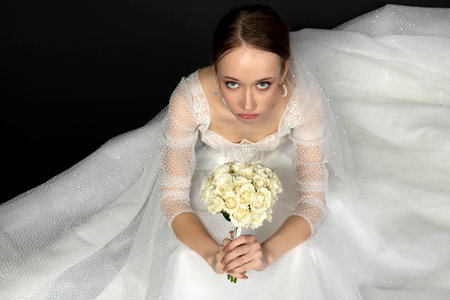 A beautiful charming girl, the bride in a luxurious and elegant white wedding dress, looking up with a graceful bouquet of white roses in her hands. Isolated on a black background. view from above.の写真素材