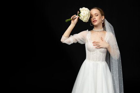 A beautiful charming girl, the bride in a luxurious and elegant white wedding dress, poses tenderly with a graceful bouquet of white roses. Isolated on a black background.の写真素材