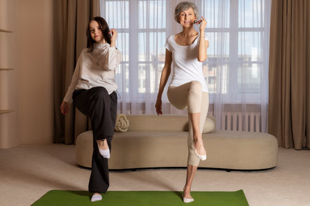 A grandmother and her granddaughter are practicing yoga in a modern beige studio. An elderly woman in excellent physical shape is teaching and sharing her experience in yoga with the young girl.の写真素材