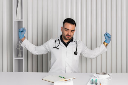 Doctor in a white coat, stethoscope on the neck and clean or sterile gloves sitting by a table doing stretches after a hard work.の写真素材