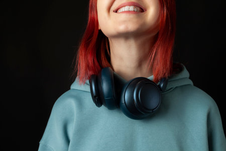 Charming young redhead girl in a blue oversized hoodie enjoying the music with a headphones. Isolated on black.の写真素材