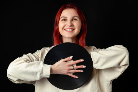Charming happy young redhead girl in a white oversized sweatshirt holds a vinyl record. Isolated on black.の写真素材