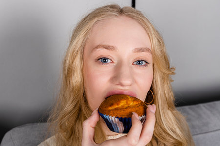 Young beautiful long haired girl in a sweater and jeans sits on a couch eating a muffin.