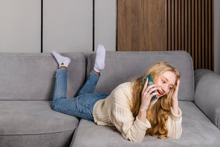 Young tender beautiful long haired girl in a sweater and jeans lies on a couch with the smart phone.の写真素材