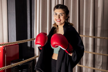 A beautiful smiling girl in red boxing gloves stands in a fighting stance and looks into the camera in a boxing ring.の写真素材
