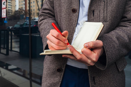 A businessman in a suit and coat is making notes in his notebook on the street, reflection of the city behind him. Business planning, Urban inspiration concept.の写真素材