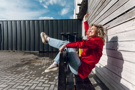 Street photography. A happy stylish curly blonde girl in red shirt posing on a street bench.の写真素材