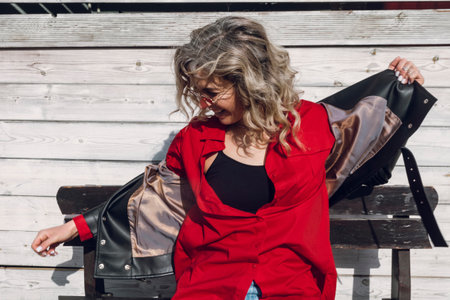 Street photography. A happy stylish curly blonde girl in red shirt posing, sitting on a bench, gray wooden planks at the background.の写真素材