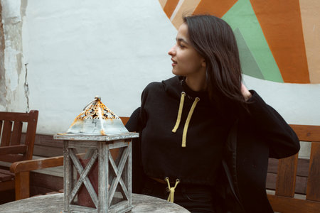 A young brunette dressed in all black posing on the street with a lantern.