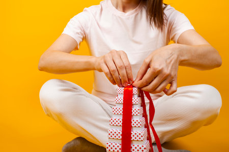 A cute brunette opening a stack of beautiful gifts or presents. unwrapping and pulling ribbons on a present. Isolated on yellow background.の写真素材