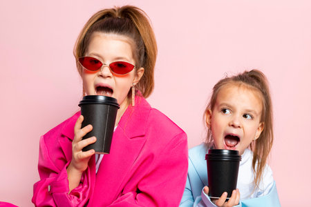 Two little girls dressed like adults. Children wearing oversized clothes. fashionable kids are holding paper cups. Isolated on pink background.の写真素材