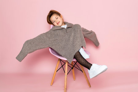Portrait of a little girl in an oversized brown sweater with a hanging sleeves. Child wearing adult clothes. Isolated on pink background.の写真素材