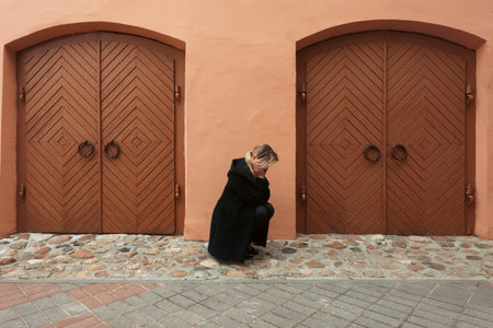 Street photography. A sad and upset young blonde girl in black is sitting alone on the street, against a beige building with two wooden doors.の写真素材