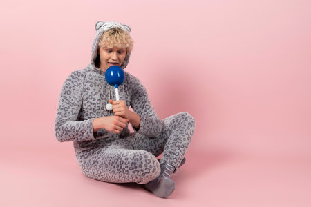 A curly blond guy in a cute and funny gray spotted animal pajamas, pretends to be a kid, holds and licks a big lollipop. Isolated on pink background.の写真素材