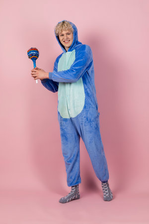 A curly blond guy in a cute and funny blue pajamas pretends to be a kid, holding a big lollipop. Isolated on pink background.の写真素材