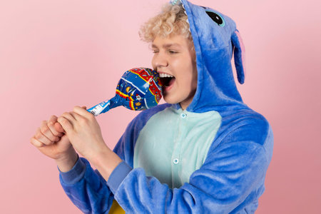 A curly blond guy in a cute and funny blue pajamas pretends to be a kid, holding a big lollipop....