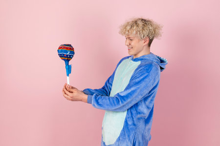A curly blond guy in a cute and funny blue pajamas pretends to be a kid, holding a big lollipop. Isolated on pink background.の写真素材