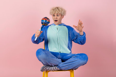 A curly blond guy in a cute and funny blue pajamas pretends to be a kid, holding a big lollipop. Isolated on pink background.の写真素材