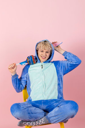 A curly blond guy in a cute and funny blue pajamas pretends to be a kid, holding a big lollipop. Isolated on pink background.の写真素材