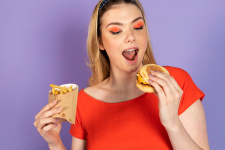Excited surprised pretty girl eats fast food, cheap burger, holding fries and burger, open mouth. fast food and unhealthy diet. isolated on purple background.の写真素材