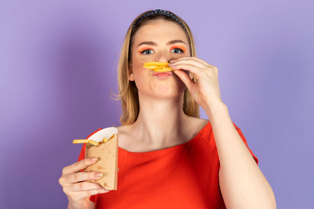 Hungry pretty girl holds fast food, oily tried potato, mimicking mustache with a fries. fast food and unhealthy diet. isolated on purple background.の写真素材