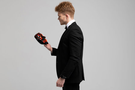 Stylish young man in a sharp black formal suit is holding a bright mask in his hands. Concealed...