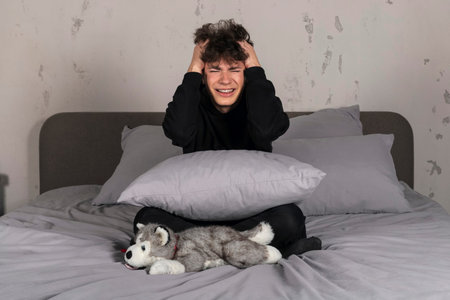 A severely depressed teenager, sitting on the bed, holding his head. A young man in a hard mental...