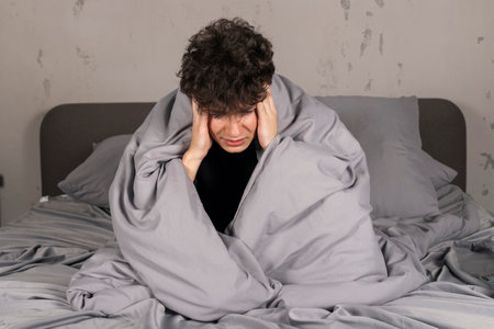 A severely depressed teenager, sits on the bed wrapped in a blanket, holds his head and crying. A young man in a hard mental condition.の写真素材