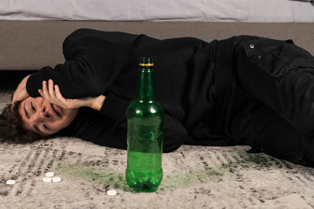 A severely depressed guy, lying on the floor among pills and a bottle of alcohol. A young man in a hard mental condition. alcohol and drugs addiction.の写真素材