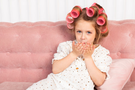 Little girl wearing hair rollers and white polka dot dress sits in the pastel pink living room, blowing a kissの写真素材