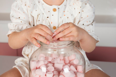 unrecognizable Little girl wearing hair rollers and white polka dot dress, sits on a pink kitchen table, with a big glass jar with sweet marshmallows or candies. puts her hands in a jar.の写真素材