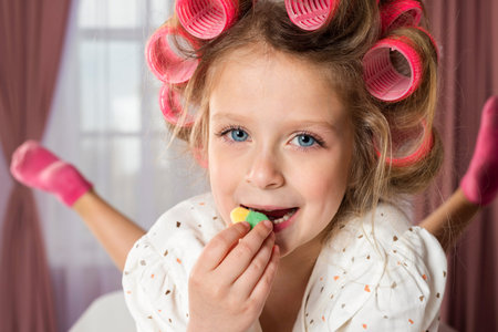 Charming little girl wearing hair rollers and white polka dot dress, eats a colorful candy.の写真素材