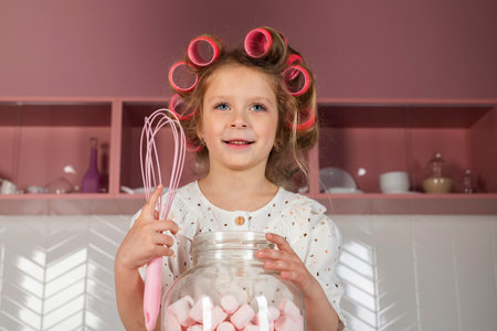 Happy Little girl wearing hair rollers and white polka dot dress, sits on a pink kitchen table, with a big glass jar with sweet marshmallows or candies.の写真素材