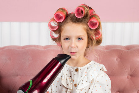 Little girl wearing hair rollers and white polka dot dress sits in the pastel pink living room, holds a hair dryer.の写真素材