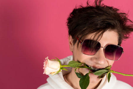 Happy, handsome young man in white hoodie, holds a rose with his teeth. A guy holding a flower with his mouth. Isolated on pink.の写真素材