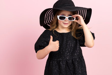 Charming little girl in black stylish summer dress, sun hat and a cool white sunglasses. Isolated on pink background.の写真素材