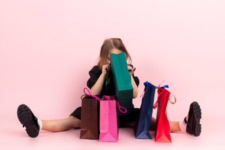 Child shopping. Charming little girl in black stylish summer dress and slight makeup sits on the floor looking in shopping bags at her new purchases. Isolated on pink background.の写真素材