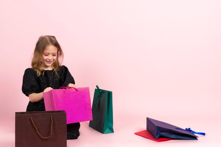 Child shopping. Charming little girl in black stylish summer dress and slight makeup sits on the floor looking in shopping bags at her new purchases. Isolated on pink background.の写真素材