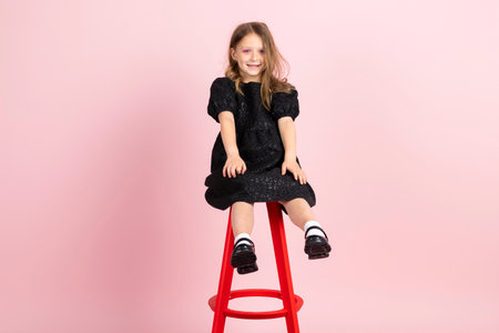Charming little girl in black stylish summer dress and slight pink makeup, sits on a tall chair. Isolated on pink background.の写真素材