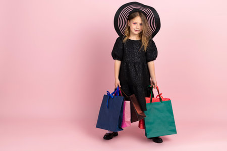 Child shopping. Charming little girl in black stylish summer dress, sun hat and slight pink makeup, posing with colorful shopping bags. Isolated on pink background.の写真素材