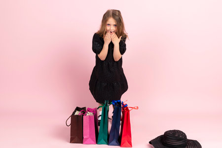 Child shopping. Charming little girl in black stylish summer dress and slight pink makeup, stands among colorful shopping bags. Isolated on pink background.の写真素材