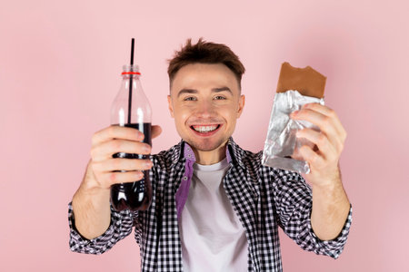 A happy young man drinking coke and eating a chocolate bar. fast food and sugar addiction, unhealthy...