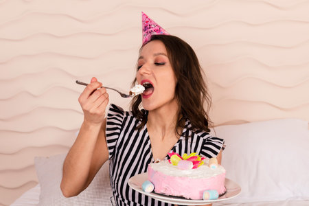 Beautiful brunette woman celebrating her birthday, dressed in a striped short dress and a birthday cap, sitting on the bed and eating a piece of the pink cakeの写真素材