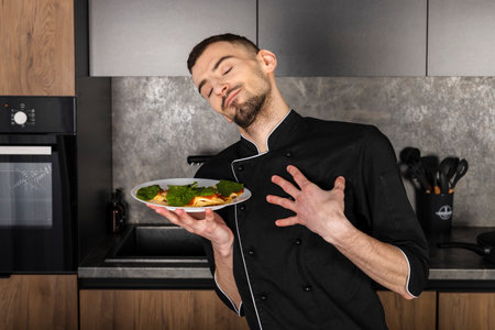 A satisfied and pleased Cook in a black uniform in the kitchen with his dish. Chef with a healthy breakfast.の写真素材