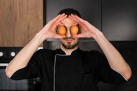 Portrait of A Cook in a black uniform in the kitchen is covering his eyes with eggs, fooling around.の写真素材